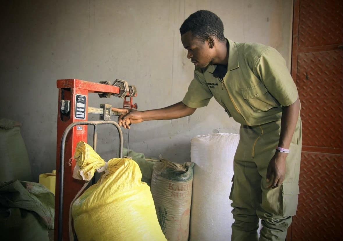 A young man in a green uniform weighs a yellow sack of produce using a manual scale at Kabubbu Development Project